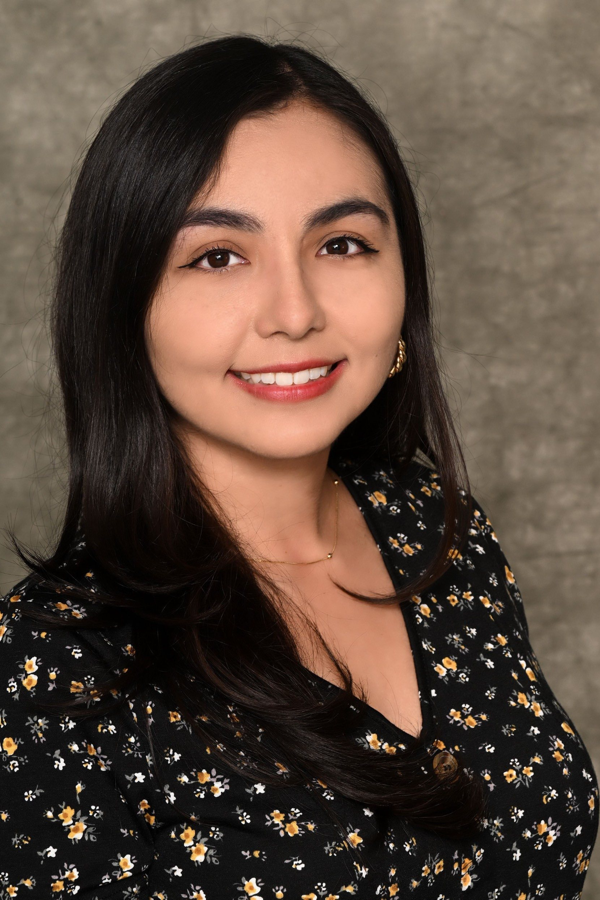 a smiling young woman, Vanessa Rodriguez, with long dark hair, wearing a black floral blouse and a gold choker