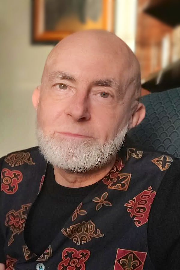 photograph of a bald man, Karl Sherlock, with a white beard, a black shirt, and a patterned vest with gold and jewel-tone designs