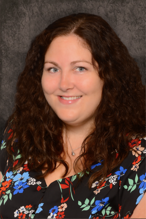 a woman, Sarah Steliga, with long curly brown hair, and wearing a black blouse with red, blue, white, and green floral patterns