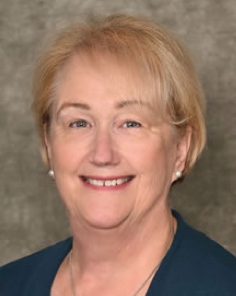 an older smiling woman with short, blonde hair, wearing a navy scoop-neck top, pearl earrings, and a pearl pendant necklace