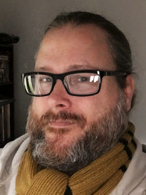 a man, Alan Traylor, with glasses, a beard, and long hair tied back, wearing a yellow and black striped scarf and a light-colored jacket