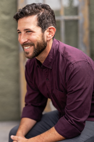 Photograph of a seated, smiling man, Adam Deutsch, with dark hair, beard, and a maroon button-up shirt.